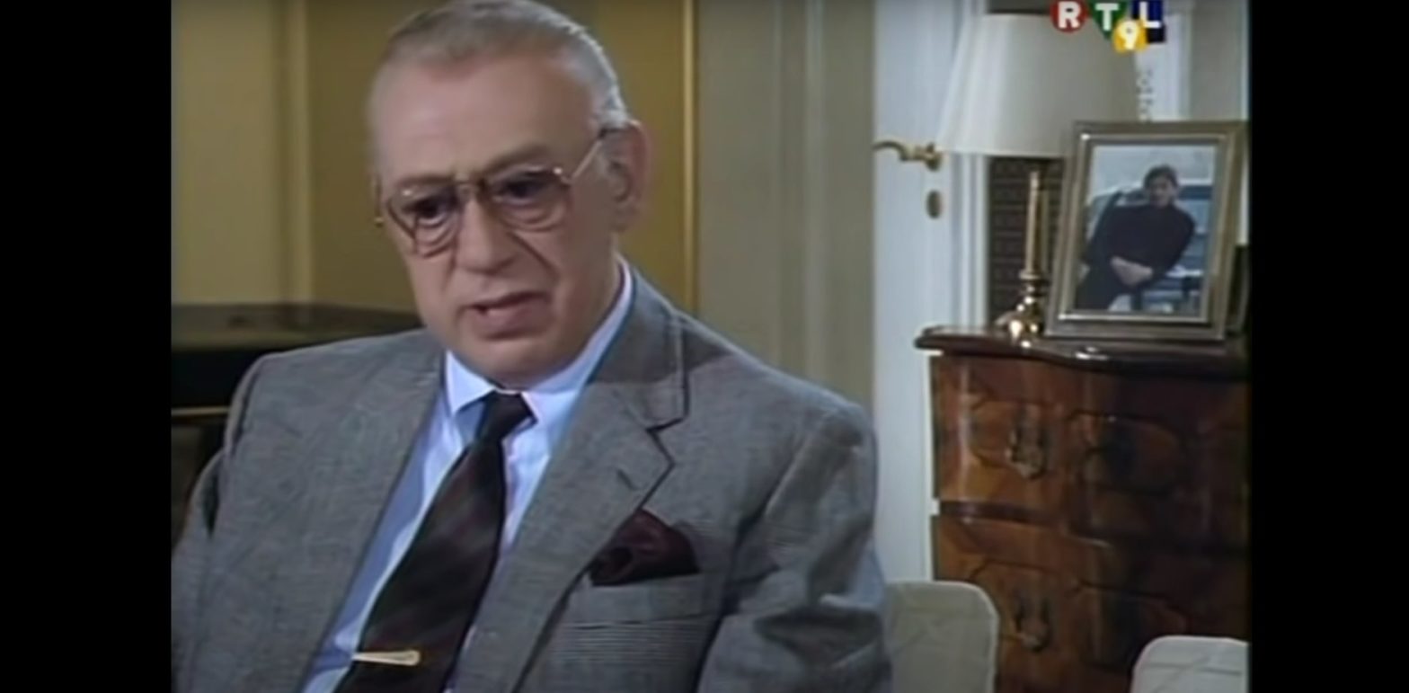 Inspecteur Derrick portant des lunettes et un costume gris assis dans un bureau classique, avec une expression sérieuse et réfléchie.